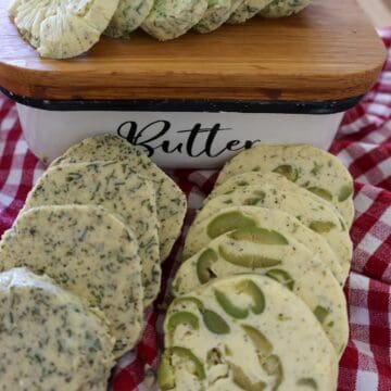 Sliced compound butter logs with herb garlic and green olive flavors next to a butter dish on a checkered towel.