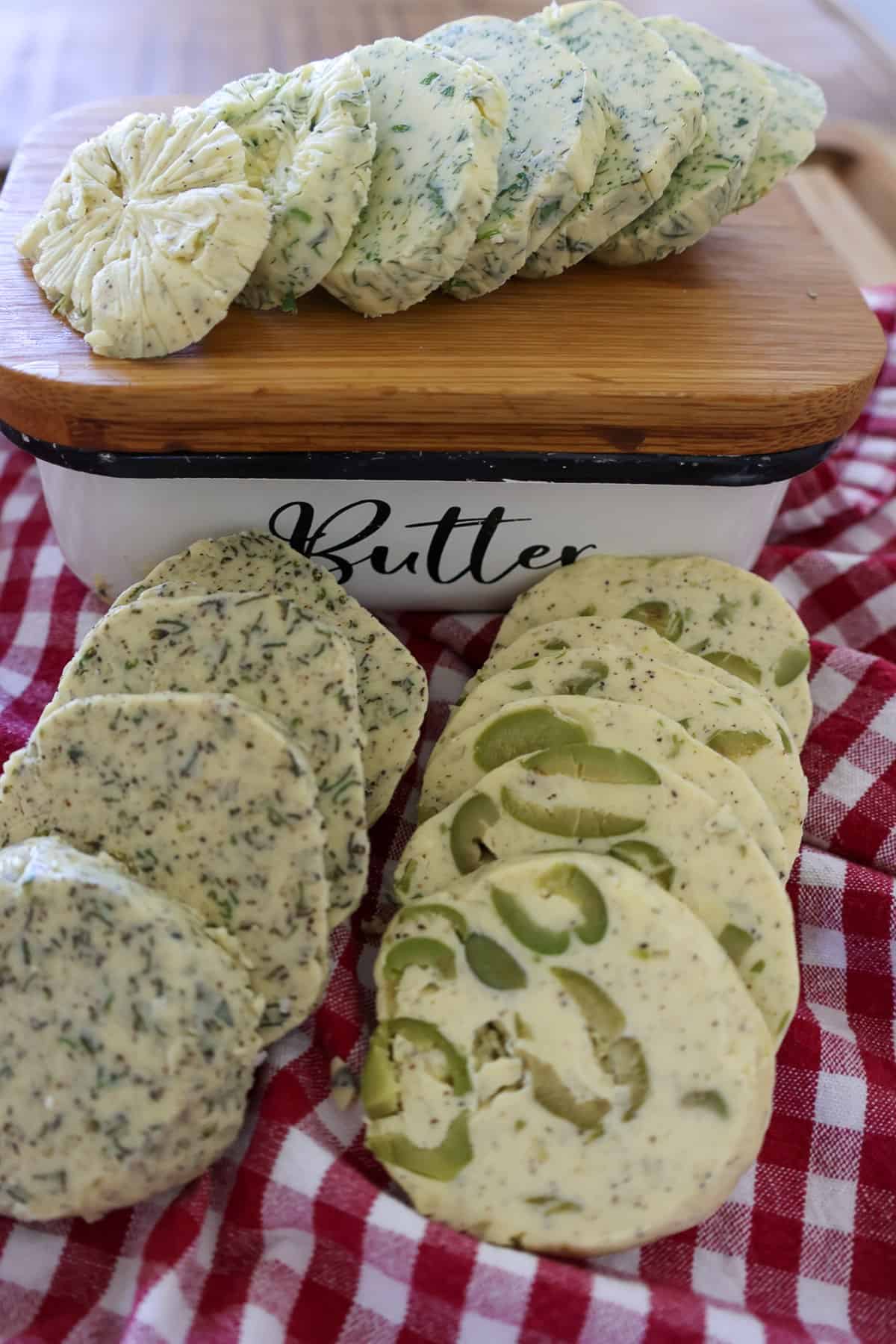 Sliced compound butter logs with herb garlic and green olive flavors next to a butter dish on a checkered towel.