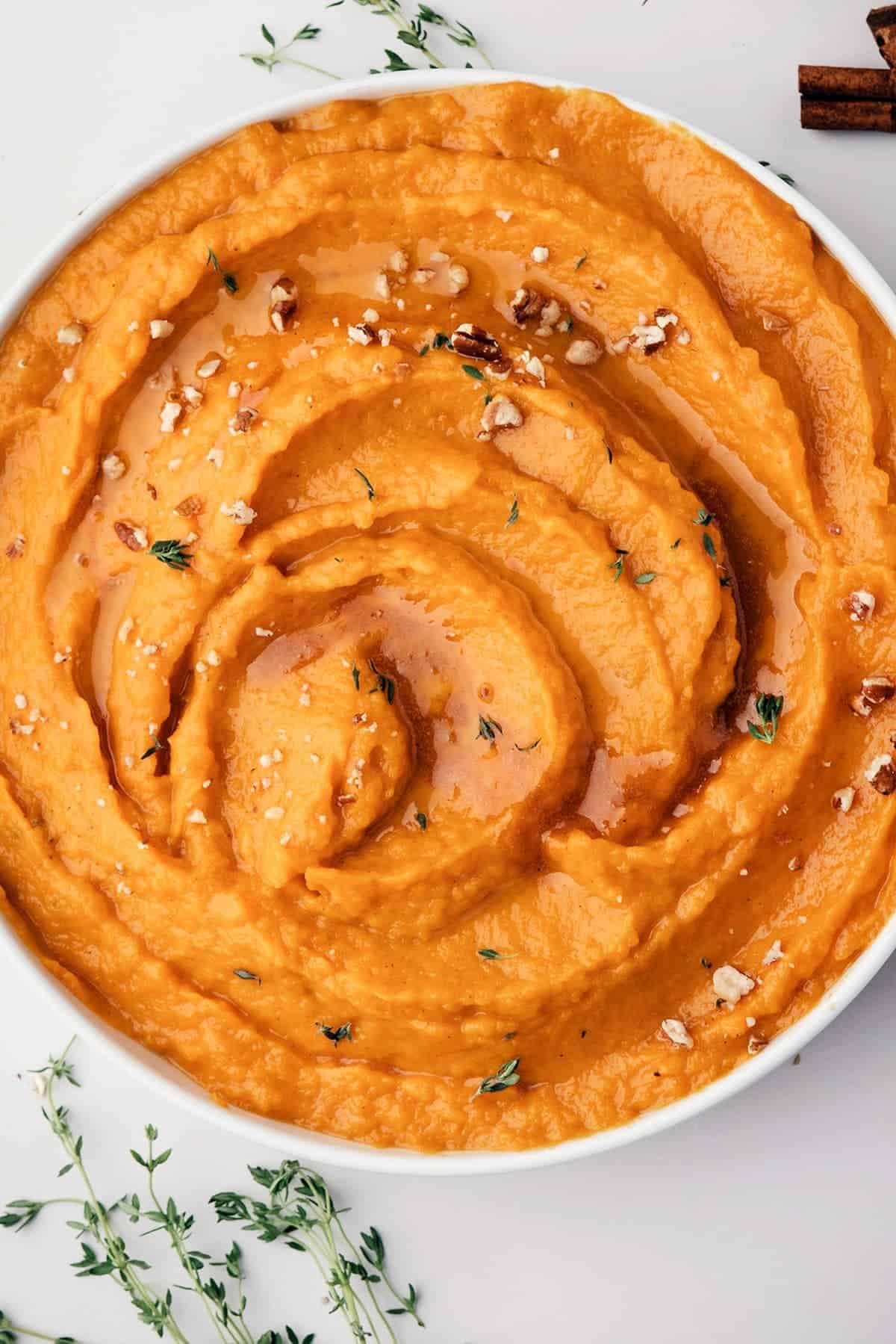 Whipped sweet potatoes swirled in a white bowl with a maple drizzle, chopped pecans, and fresh thyme.