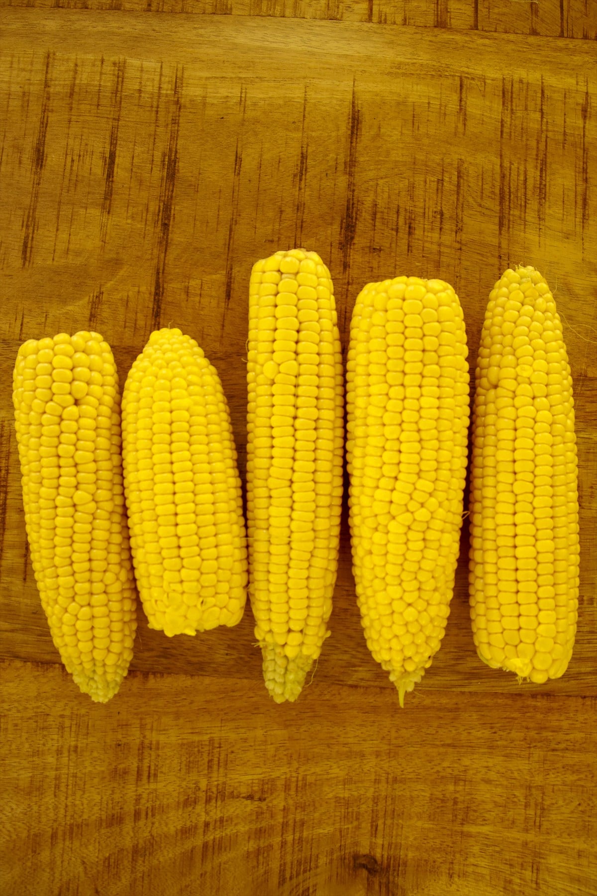 Five husked ears of corn trimmed flat on a wooden cutting board for corn ribs.