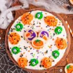 Halloween sugar cookie cheesecake decorated with orange and green frosting swirls, candy eyes, sprinkles, and pumpkin and ghost sugar cookies on a wooden board.