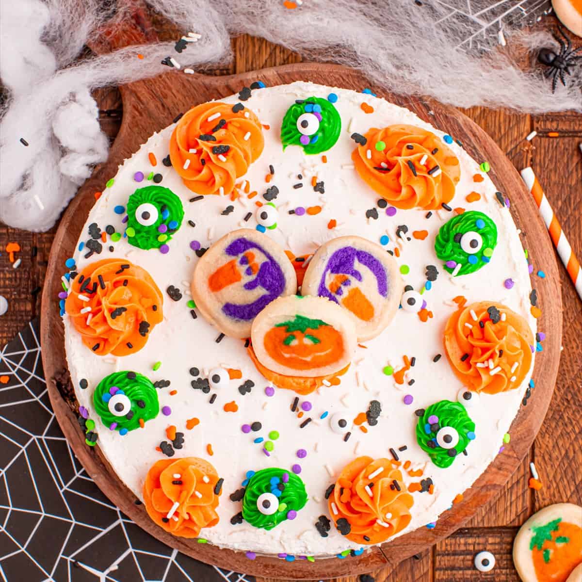 Halloween sugar cookie cheesecake decorated with orange and green frosting swirls, candy eyes, sprinkles, and pumpkin and ghost sugar cookies on a wooden board.