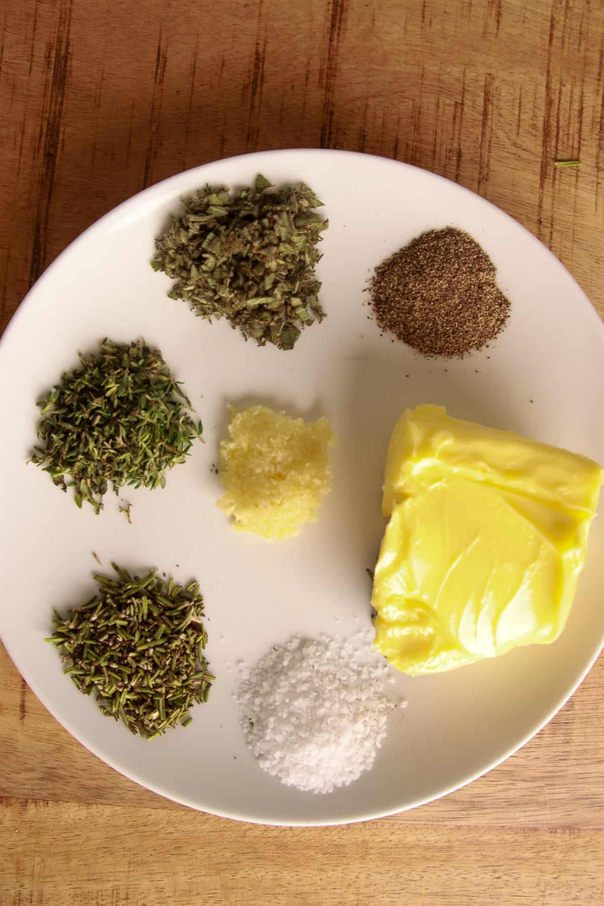 Softened butter on a plate with chopped thyme, rosemary, sage, grated garlic, salt, and pepper for herb and garlic compound butter.