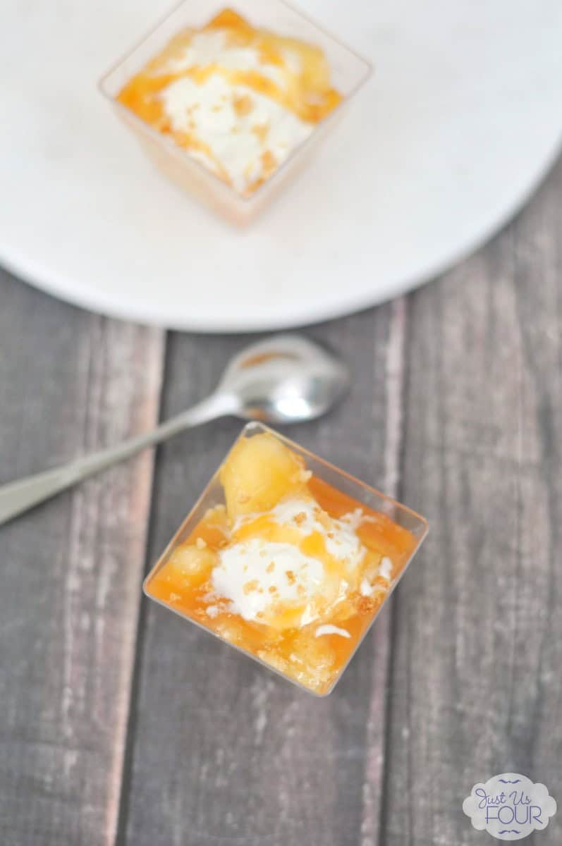 Two small square glasses layered with spiced apple pie filling, caramel sauce, whipped cream and crumbled pie crust, one on a white plate and one on a wood table with a spoon.