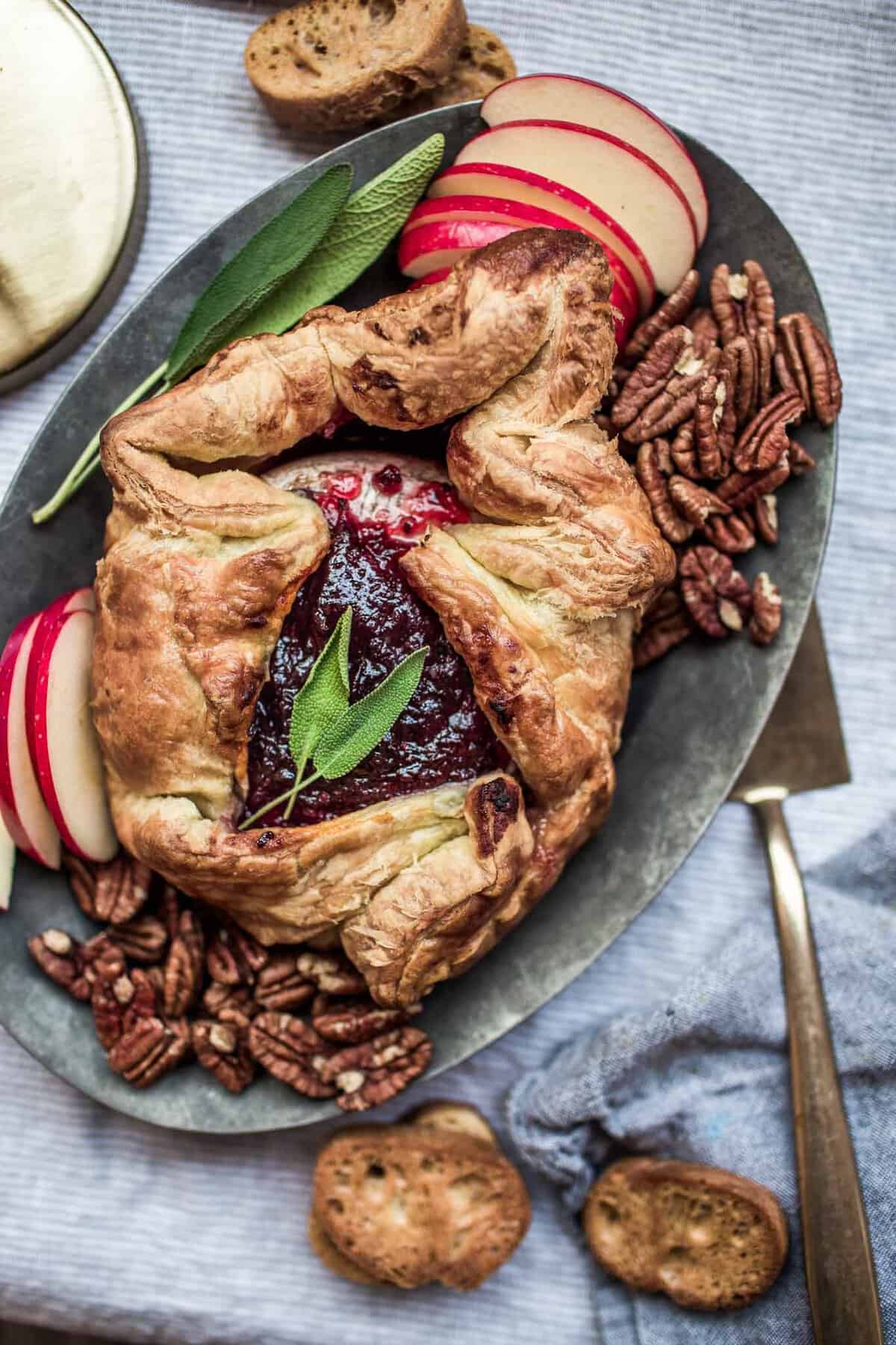 Golden baked brie wrapped in puff pastry with a jam center and sage, on a platter with apple slices and pecans.