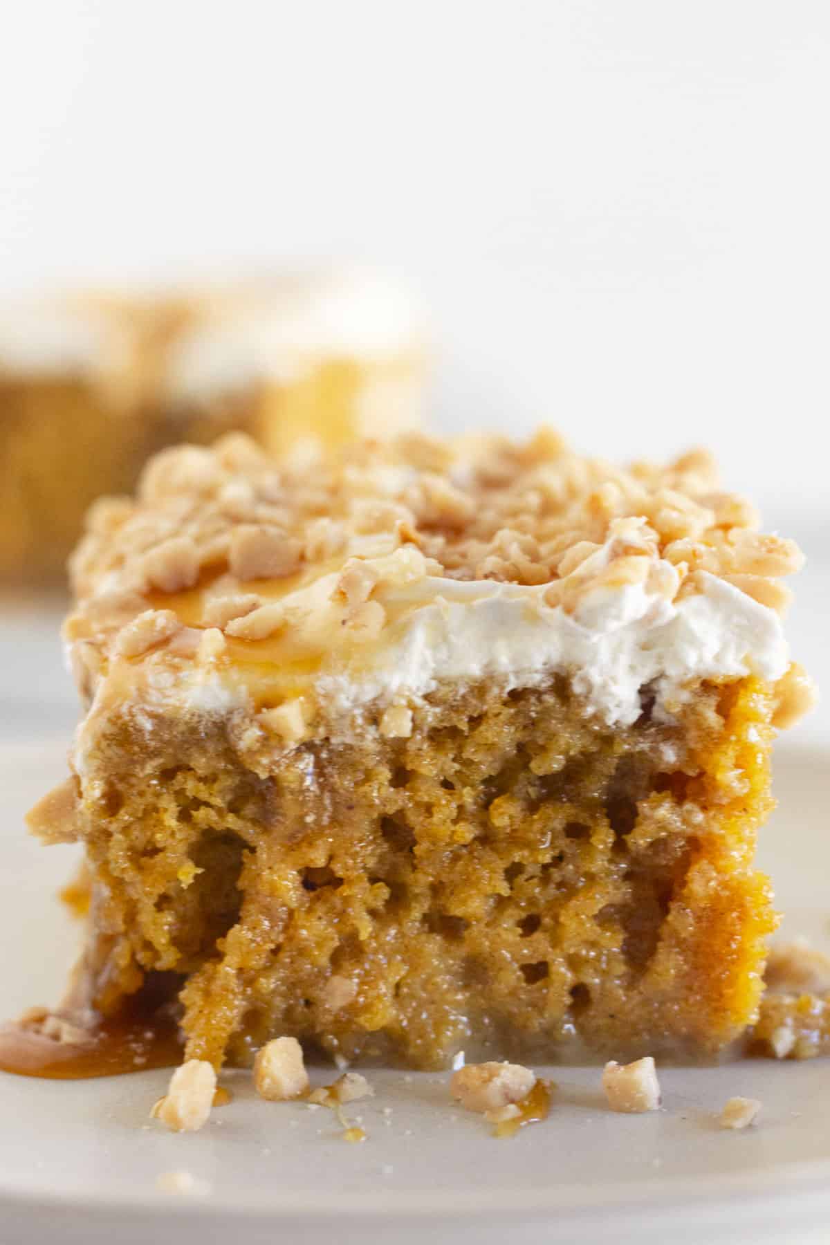 A slice of pumpkin poke cake with caramel, whipped topping, and toffee bits on a white plate.