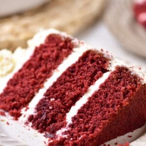 A slice of red velvet cake with three deep red layers and mascarpone frosting on a white plate.