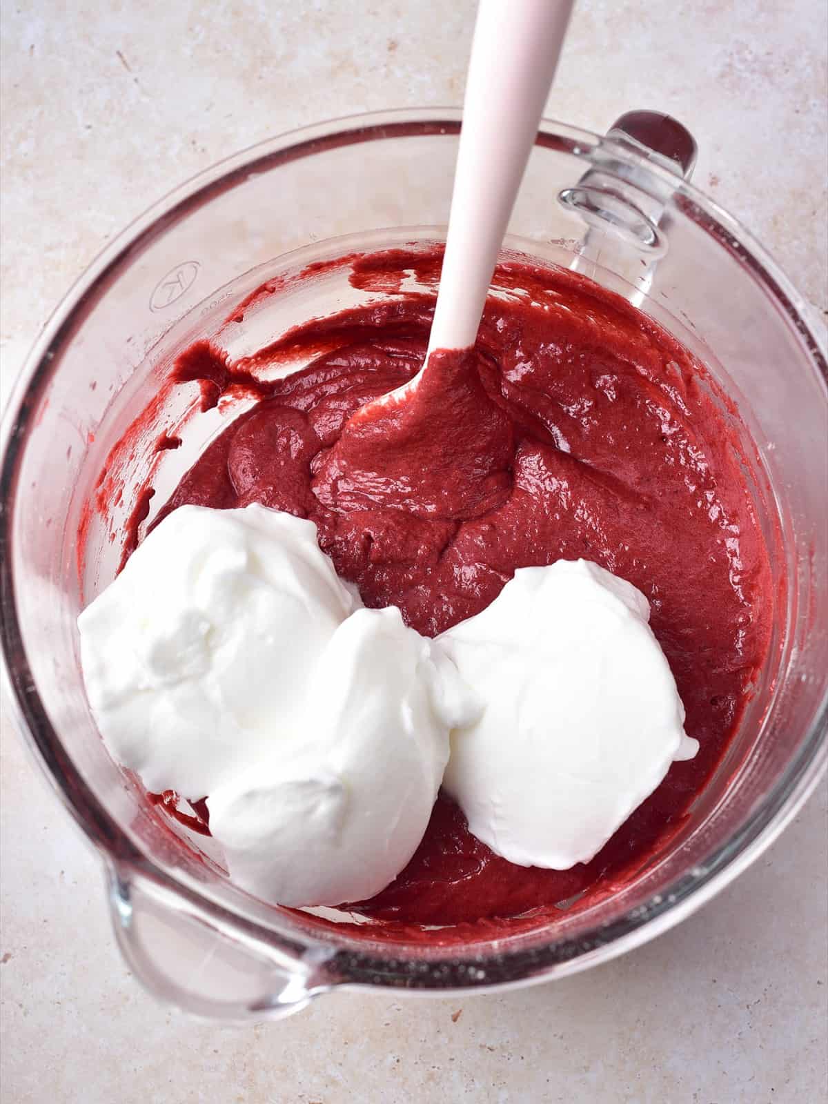 Stiffly beaten egg whites being folded into the red velvet cake batter with a pink spatula.