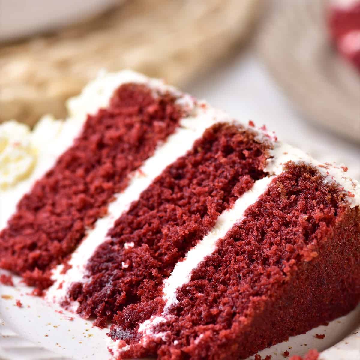 A slice of red velvet cake with three deep red layers and mascarpone frosting on a white plate.