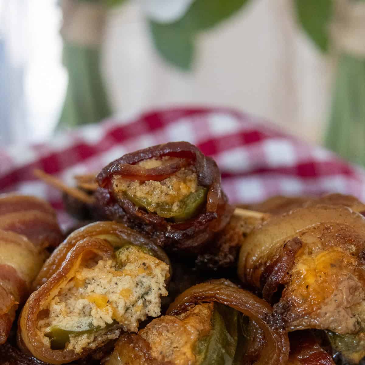 Smoked bacon wrapped jalapeno poppers piled on a plate with a red gingham napkin behind them.