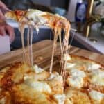Smoked pizza with three cheeses being served with a gooey cheese pull from a slice lifted off the cutting board.