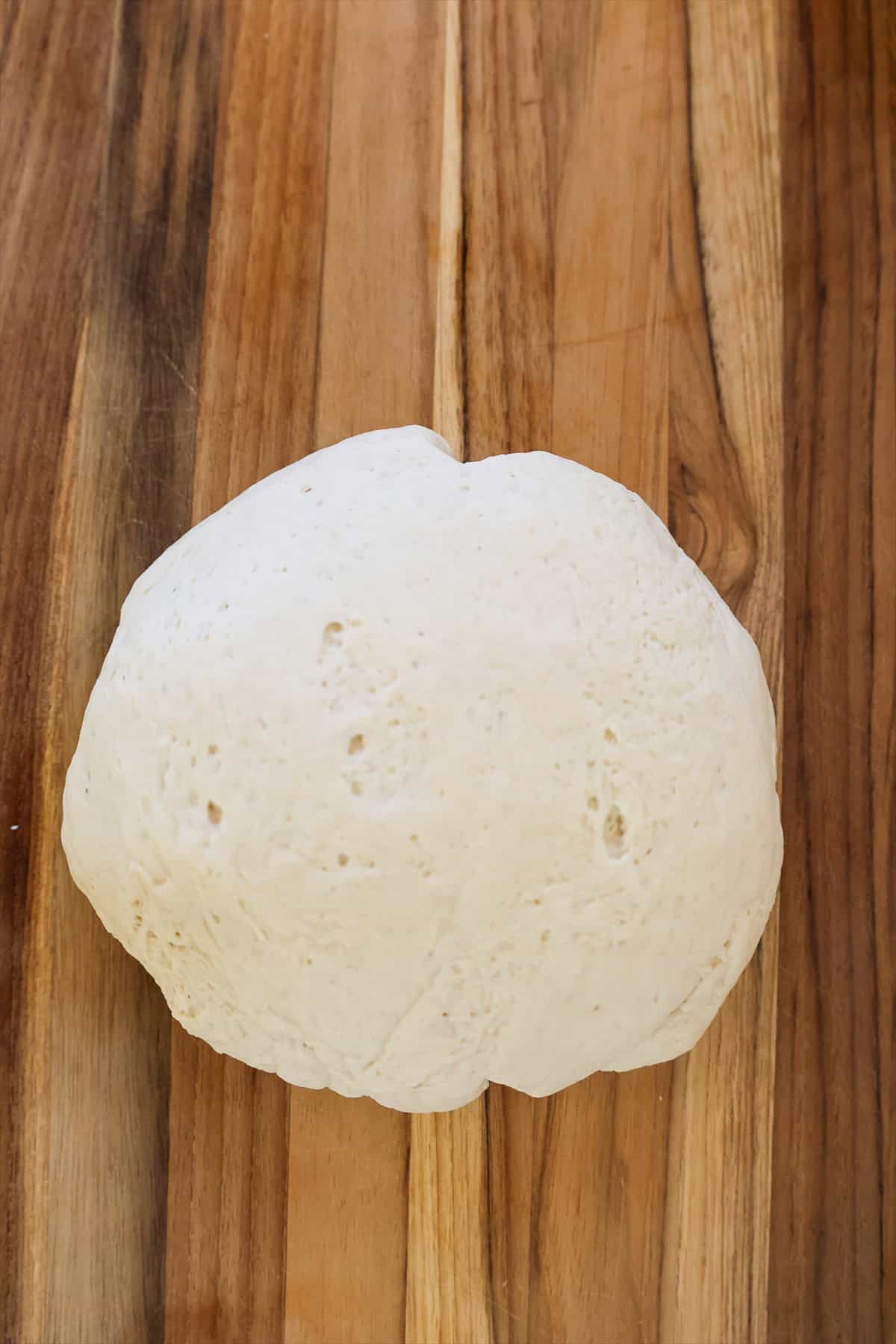 Ball of room temperature pizza dough resting on a wooden cutting board.