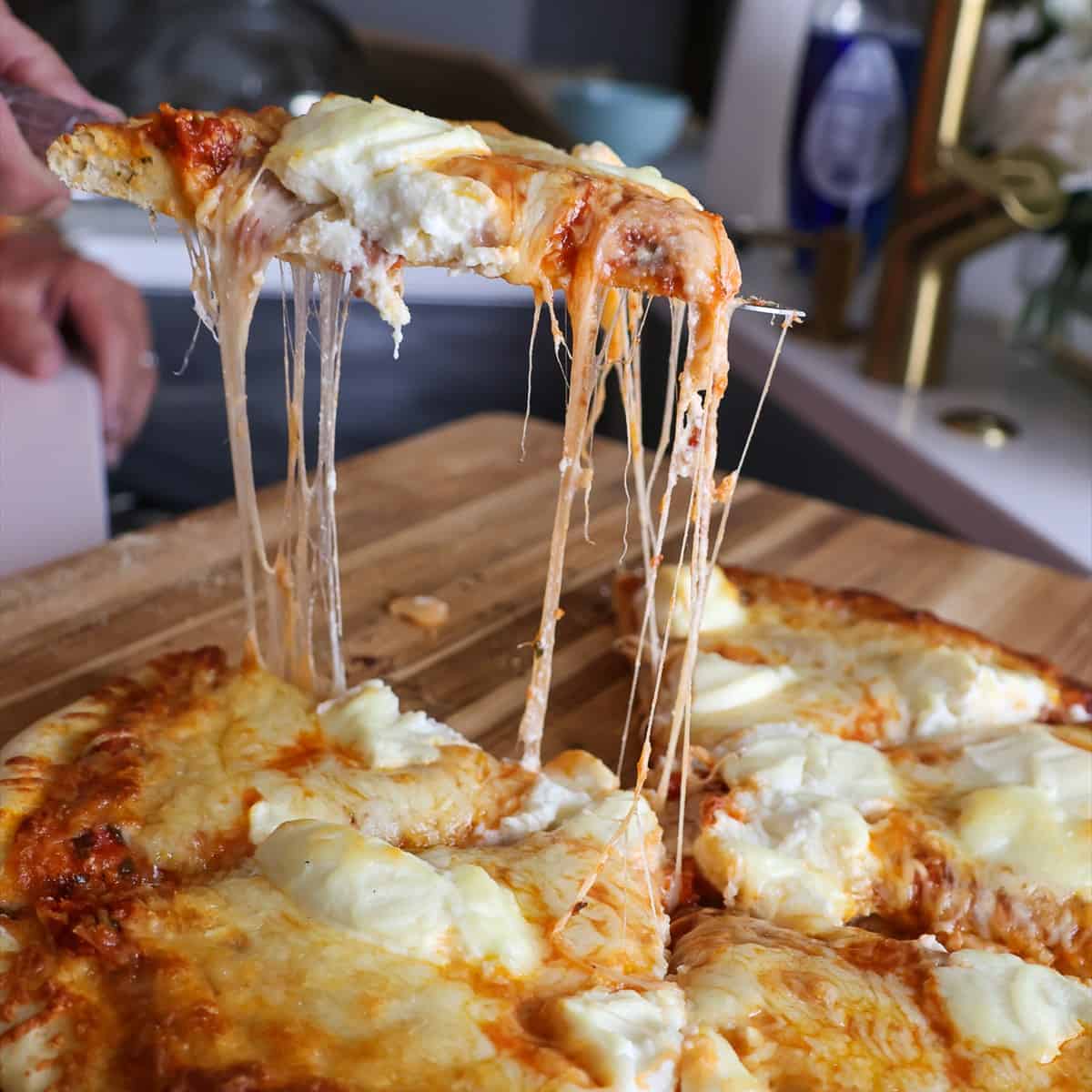 Smoked pizza with three cheeses being served with a gooey cheese pull from a slice lifted off the cutting board.