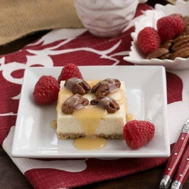 A caramel pecan cheesecake bar drizzled with caramel and topped with pecans, with raspberries on the plate.