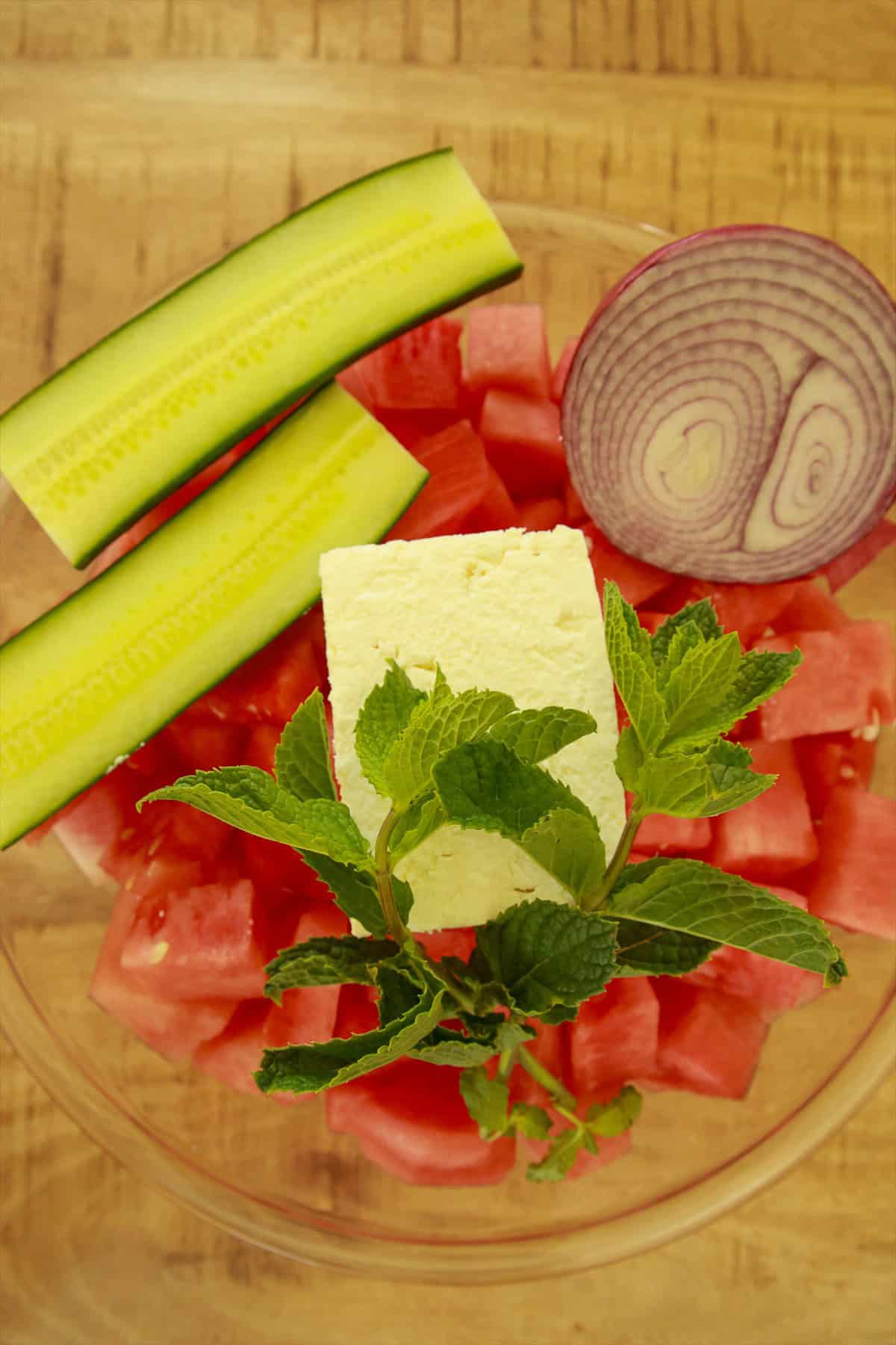 Watermelon cubes, cucumber, feta, mint, and red onion layered in a large glass bowl for watermelon feta salad.