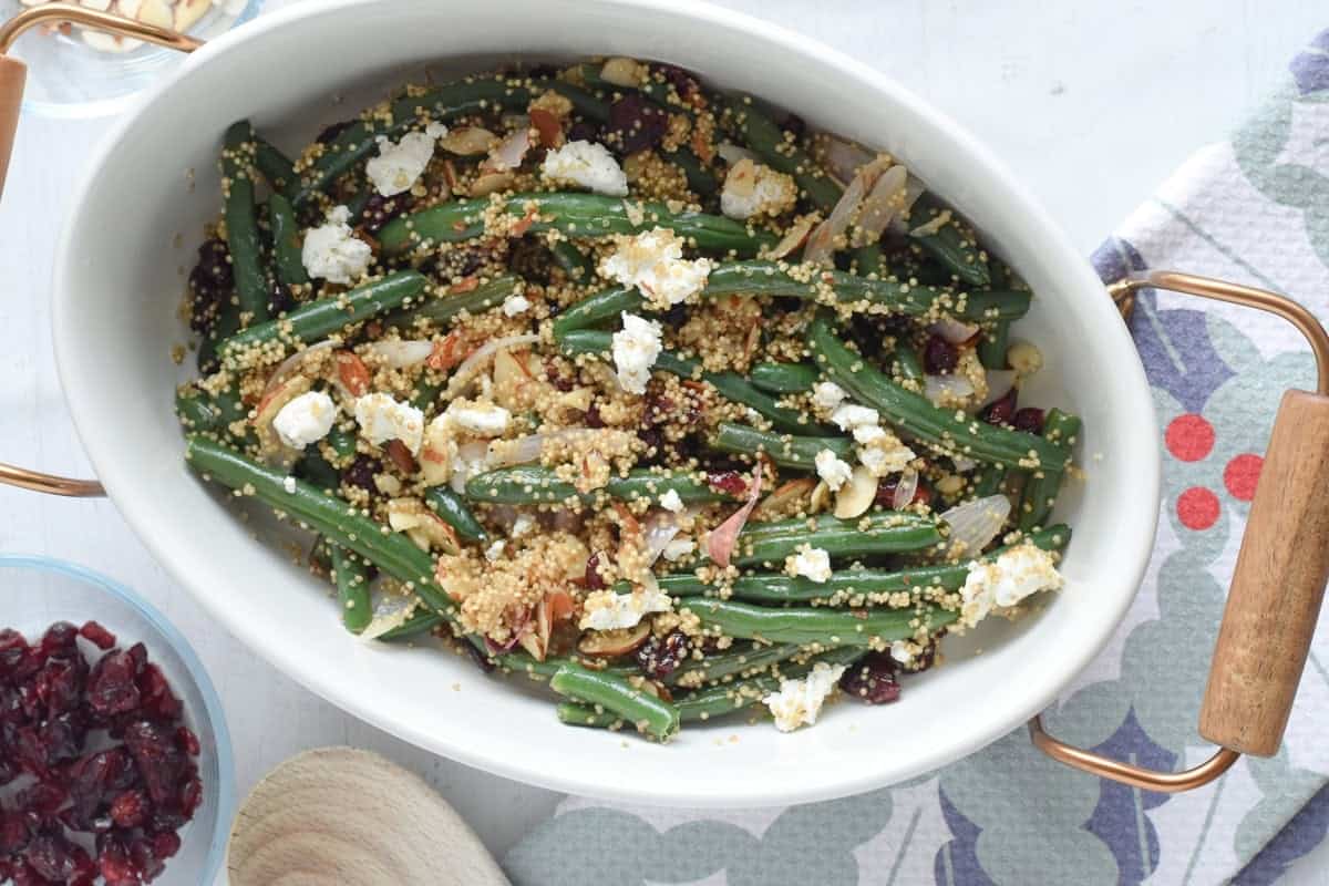 Cranberry green beans with quinoa, sliced almonds, shallots, and crumbled goat cheese in a white oval baking dish.