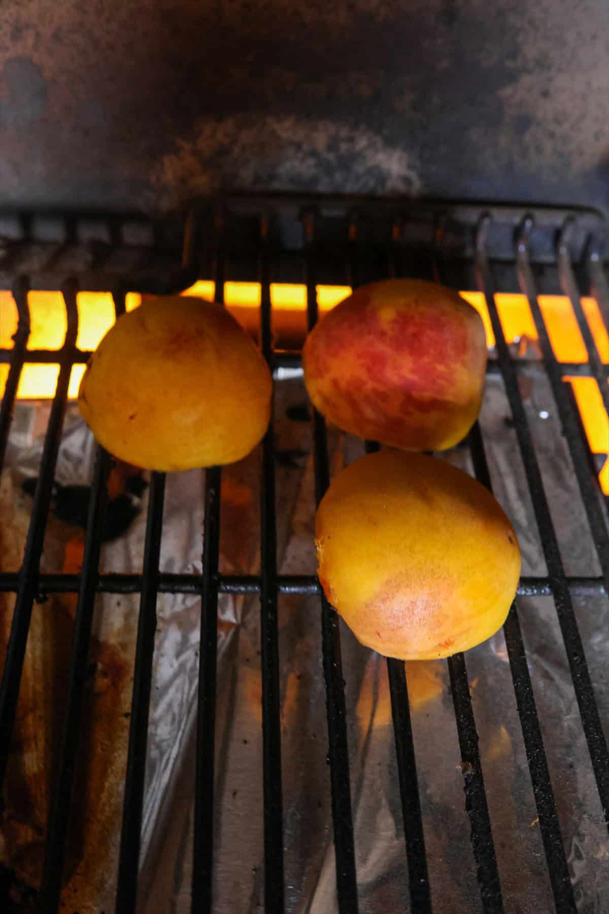 Buttered peach halves cooking on hot grill grates over glowing heat.