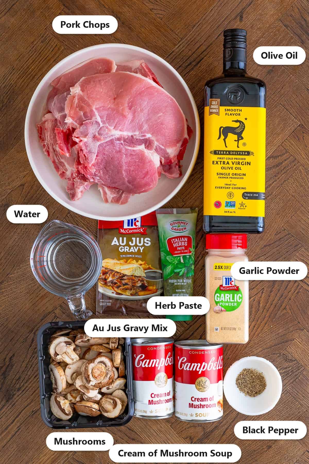 Labeled ingredients for slow cooker pork chops including bone in pork chops, olive oil, mushrooms, cream of mushroom soup, au jus gravy mix, herb paste, garlic powder, black pepper, and water.