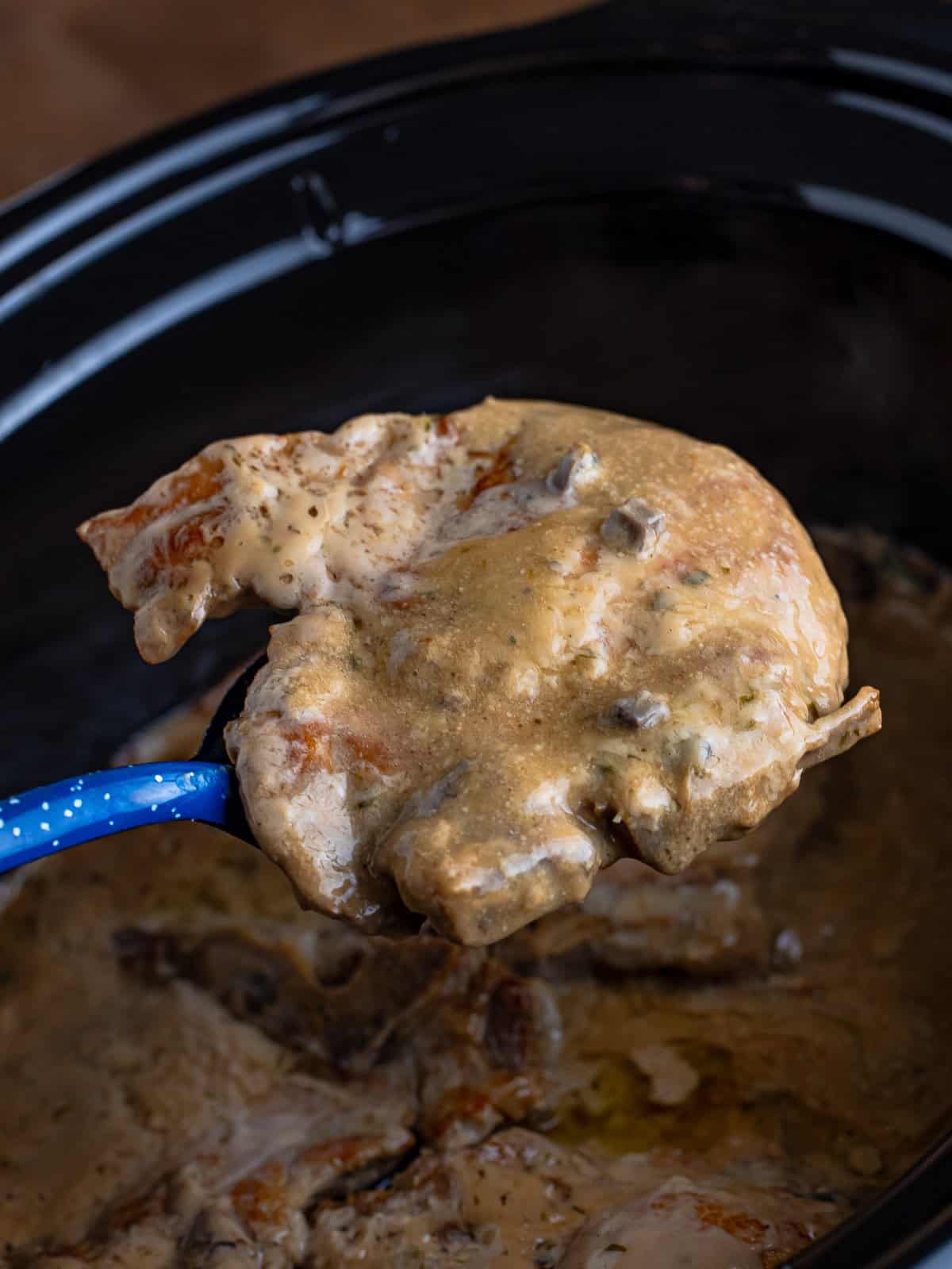 A serving spoon lifting a tender pork chop with mushroom gravy out of the slow cooker.