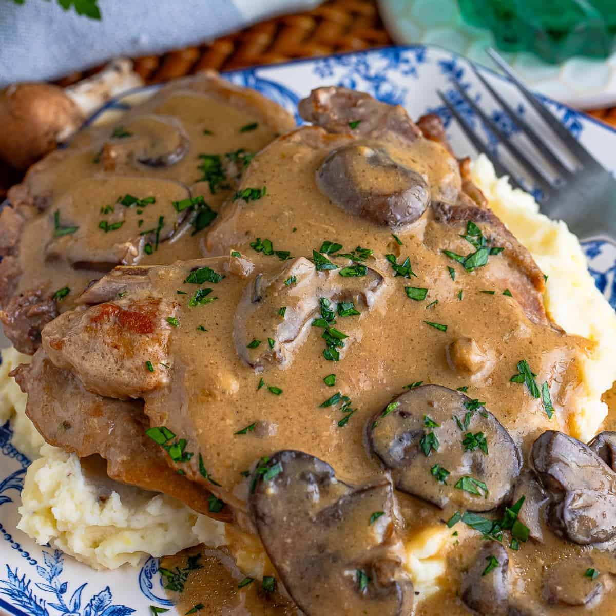 Slow cooker pork chops smothered in creamy mushroom gravy over mashed potatoes on a blue and white plate.