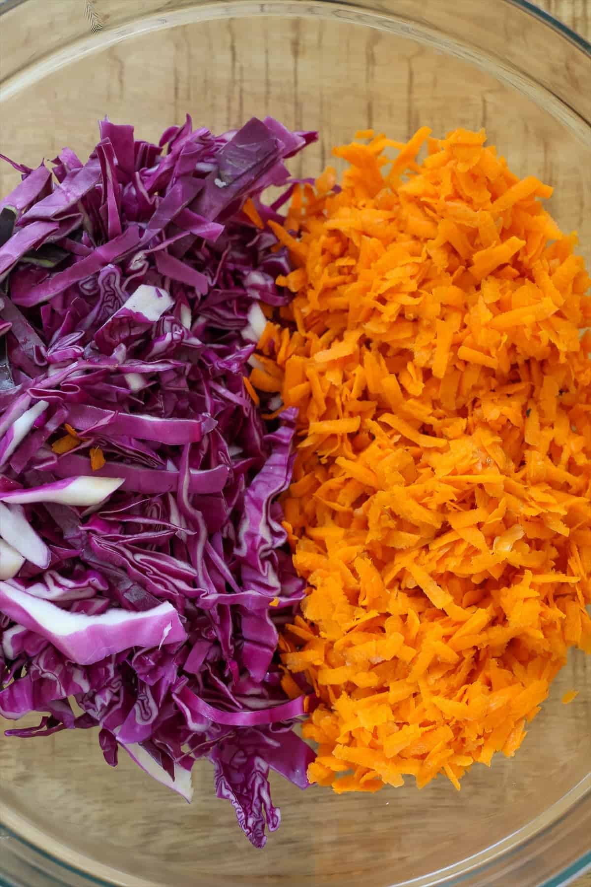 Shredded red cabbage and grated carrots side by side in a large glass mixing bowl.